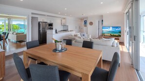 A modern kitchen with white cabinets, a stainless steel refrigerator, and a dining area with a wooden table and grey chairs. - Oasis 21, Oasis 21 on Hamilton island (Hamilton Island)