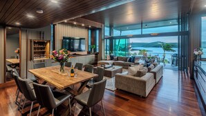 A spacious living area with a wooden dining table, comfortable seating, and a large window offering a view of the sea. - Yacht Club Villa 29, Yacht Club Villa 29 on Hamilton Island (Hamilton Island)