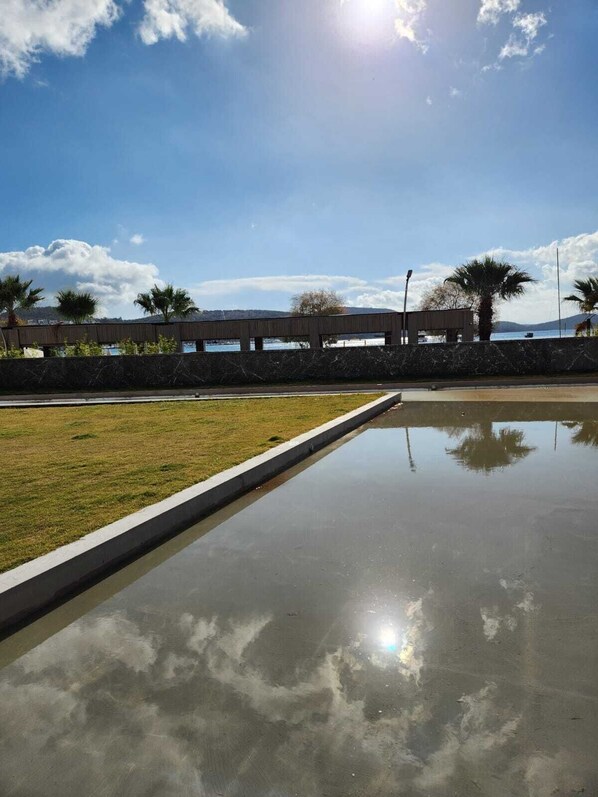 A paved area with a water feature reflecting the sky, palm trees, and a distant view of a body of water and hills. - SEA SOUL OTEL (Bodrum)