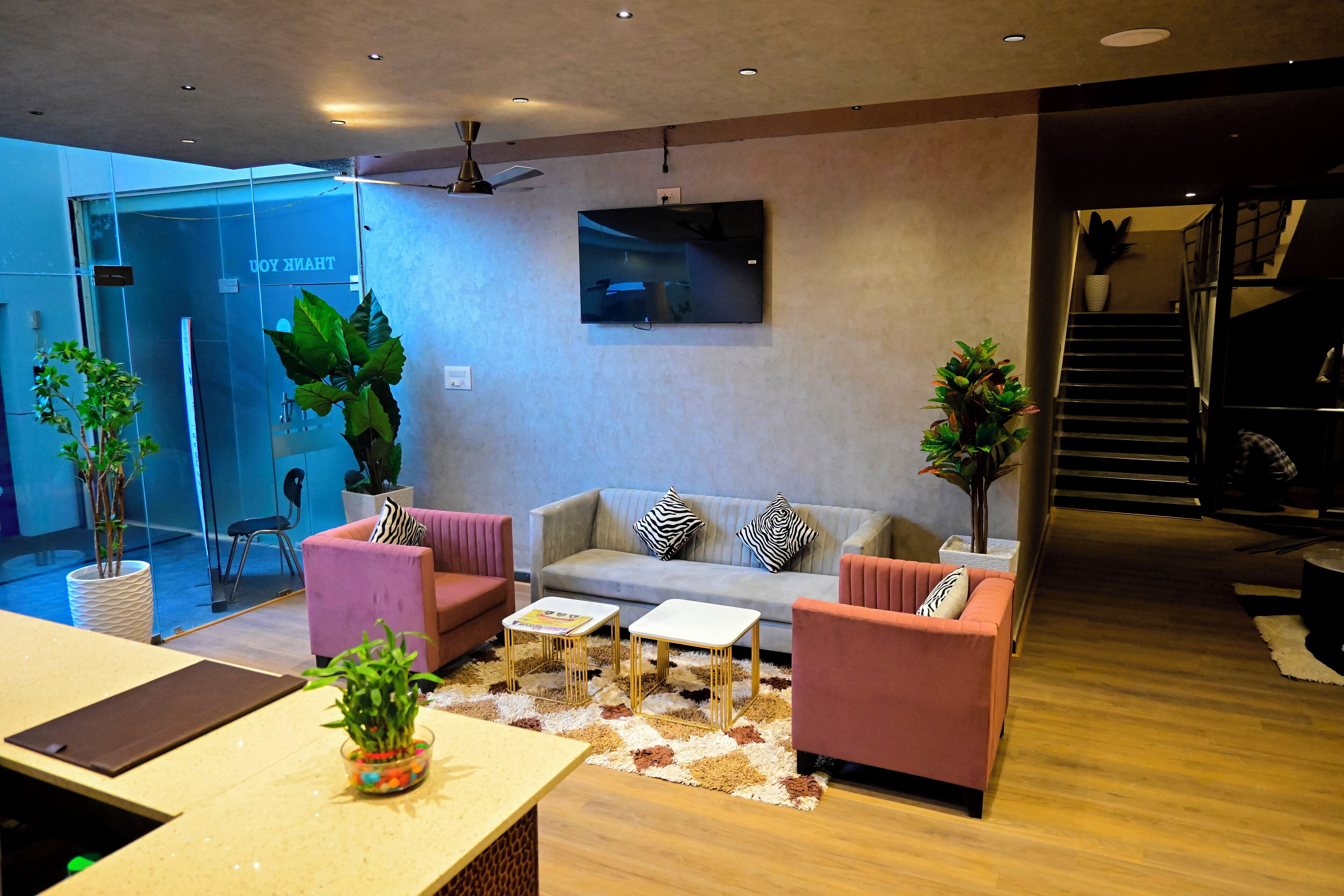 A modern living room with a sofa, two armchairs, a coffee table, and a TV mounted on the wall. There are potted plants and a staircase leading to an upper level.