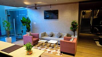 A modern living room with a sofa, two armchairs, a coffee table, and a TV mounted on the wall. There are potted plants and a staircase leading to an upper level.