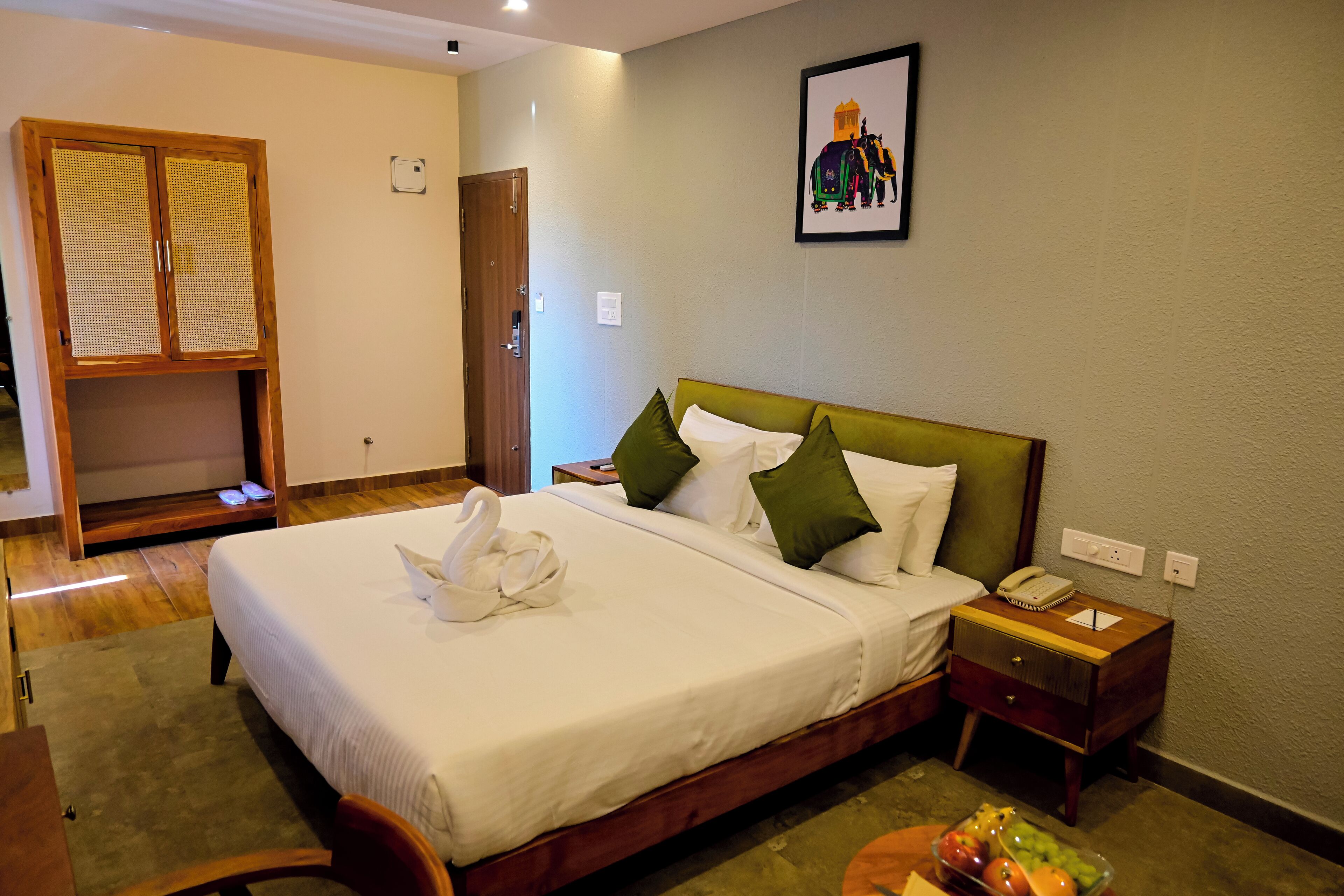 A neatly arranged hotel room with a large bed, a wooden wardrobe, a bedside table with a lamp, and a small table with fruit.