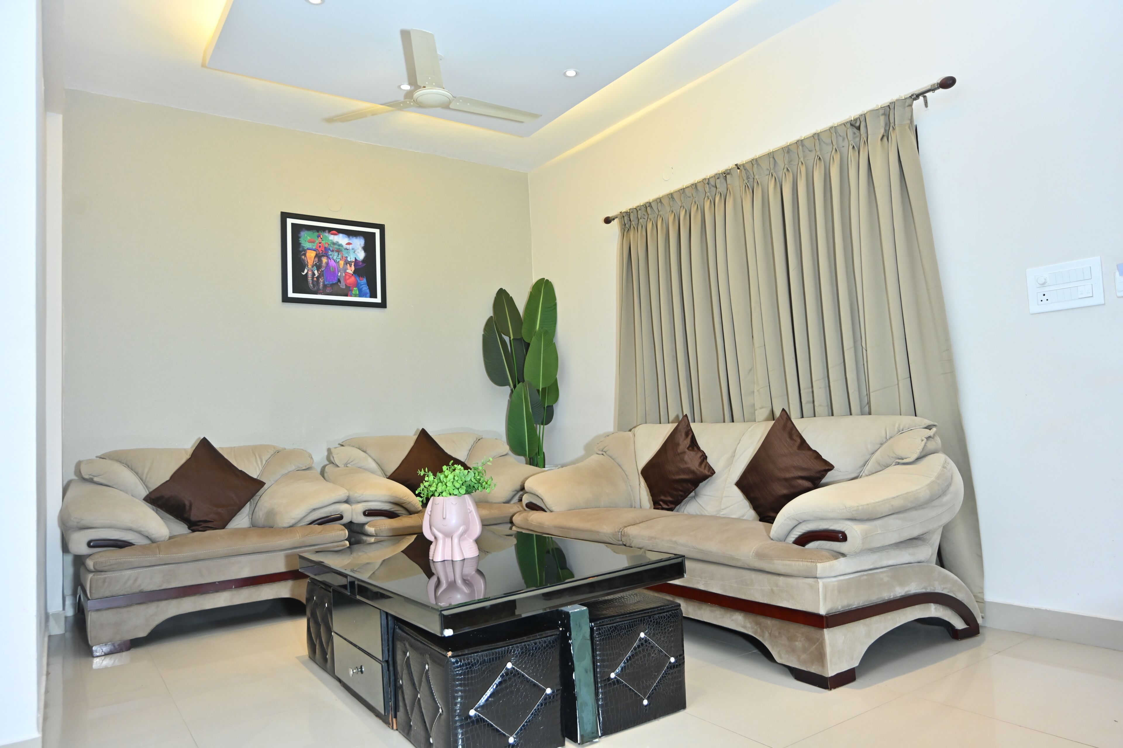 A living room with two sofas, a coffee table, a ceiling fan, a large window with curtains, and a wall-mounted picture.
