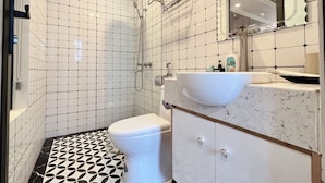 Casa de banho com sanita branca, lavatório com bancada de mármore e zona de duche com porta de vidro.