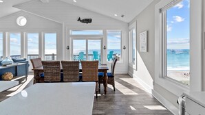 A spacious dining area with a large table and wicker chairs, large windows with ocean views, and a ceiling with recessed lighting. - In Spiaggia (at the beach) #185 - 6 BD Oceanfront in the heart of Surf City! (Surf City)