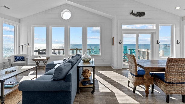 A spacious living area with a large window overlooking the sea, a cozy seating area with a sofa and chairs, and a wooden dining table. - In Spiaggia (at the beach) #185 - 6 BD Oceanfront in the heart of Surf City! (Surf City)