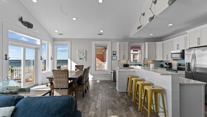 A modern kitchen with white cabinets, a central island with stools, and a dining area with wicker chairs. - In Spiaggia (at the beach) #185 - 6 BD Oceanfront in the heart of Surf City! (Surf City)
