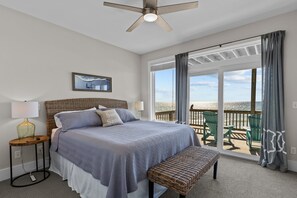 A bedroom with a bed, bedside table, lamp, and a view of the beach through a large window. - In Spiaggia (at the beach) #185 - 6 BD Oceanfront in the heart of Surf City! (Surf City)