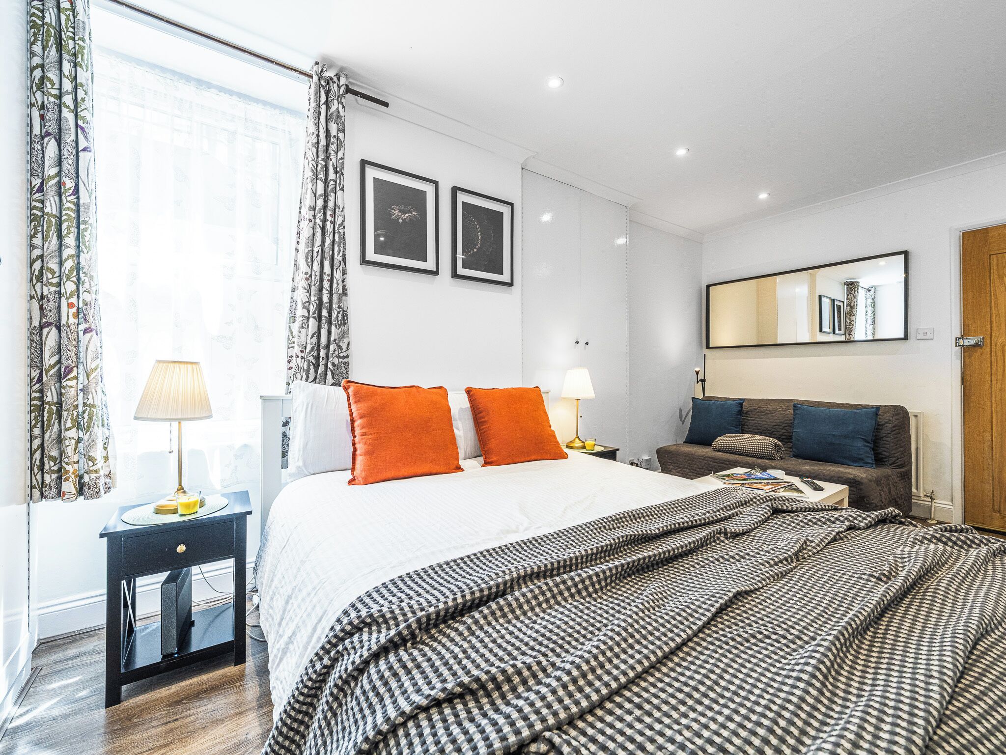 A modern hotel room with a large bed, two bedside tables, a sofa, a mirror, and framed pictures on the wall.