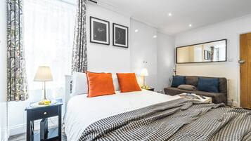 A modern hotel room with a large bed, two bedside tables, a sofa, a mirror, and framed pictures on the wall.