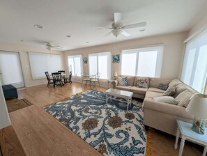 Uma sala de estar espaçosa com uma janela grande, um ventilador de teto, um sofá, uma mesa de centro e um espaço de jantar com cadeiras.