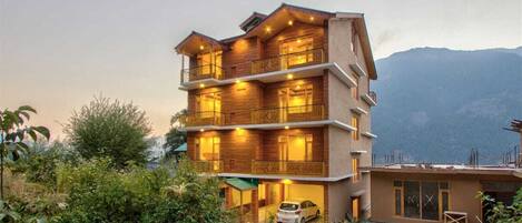 A multi-story hotel with balconies, a car parked in front, and a mountainous backdrop.