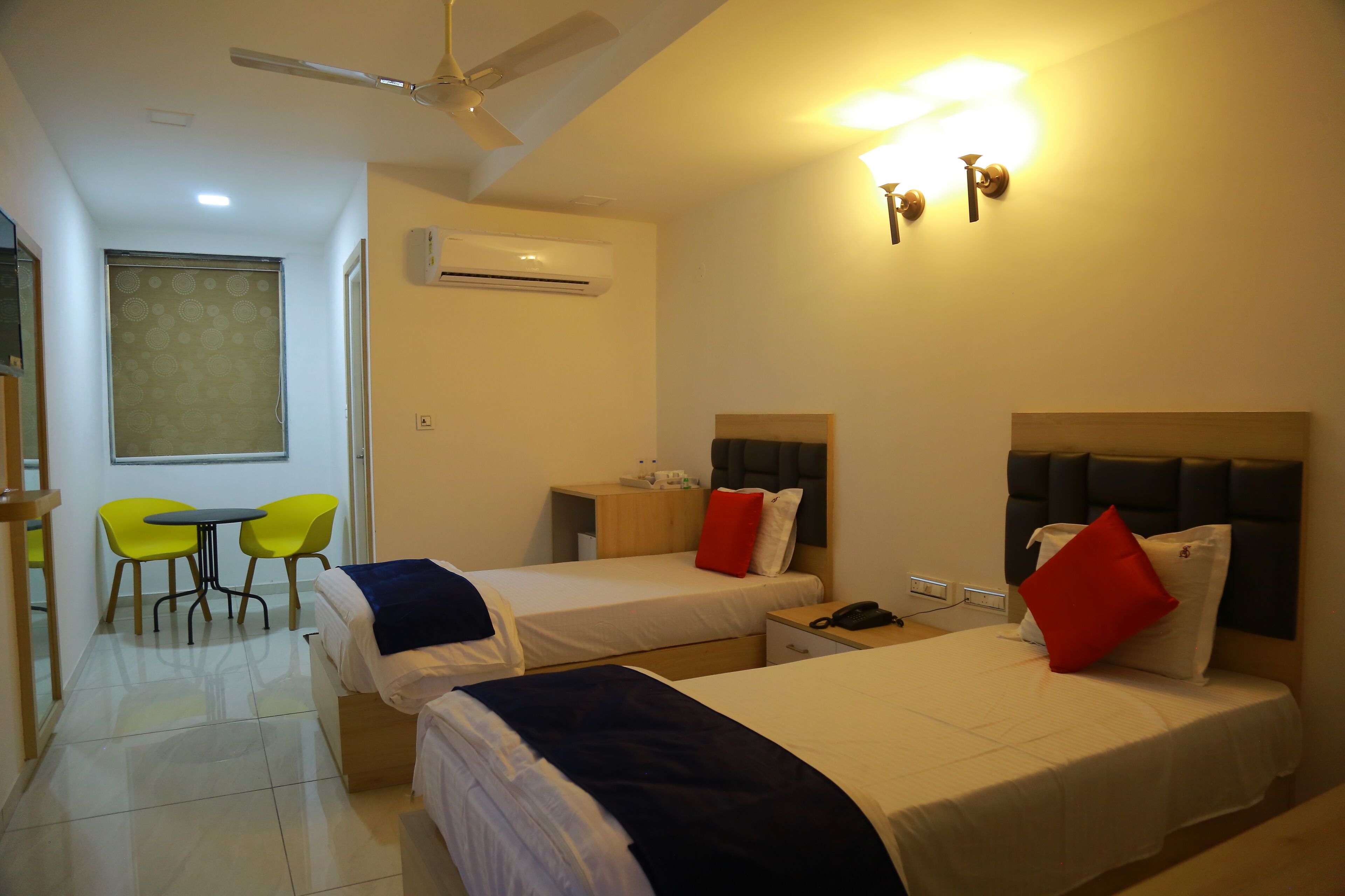 A hotel room with two beds, a small table with chairs, an air conditioning unit, and a ceiling fan.
