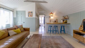 Amplia sala de estar con un sofá de cuero marrón, una mesa de centro de madera y un tapete estampado. Hay dos taburetes azules cerca de una cocina con encimera de madera y una guitarra.
