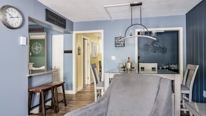 A living room with a grey armchair, a dining table with chairs, a wall clock, and a motorcycle decoration.