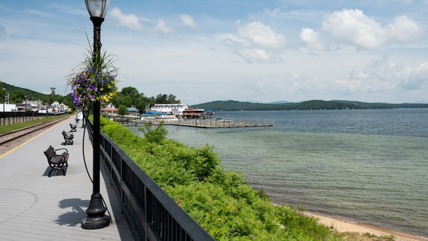 A waterfront promenade with benches and a lamp post, overlooking a calm lake and distant buildings.