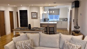 Una sala de estar moderna con un sofá seccional beige grande, piso de madera y una cocina al fondo.