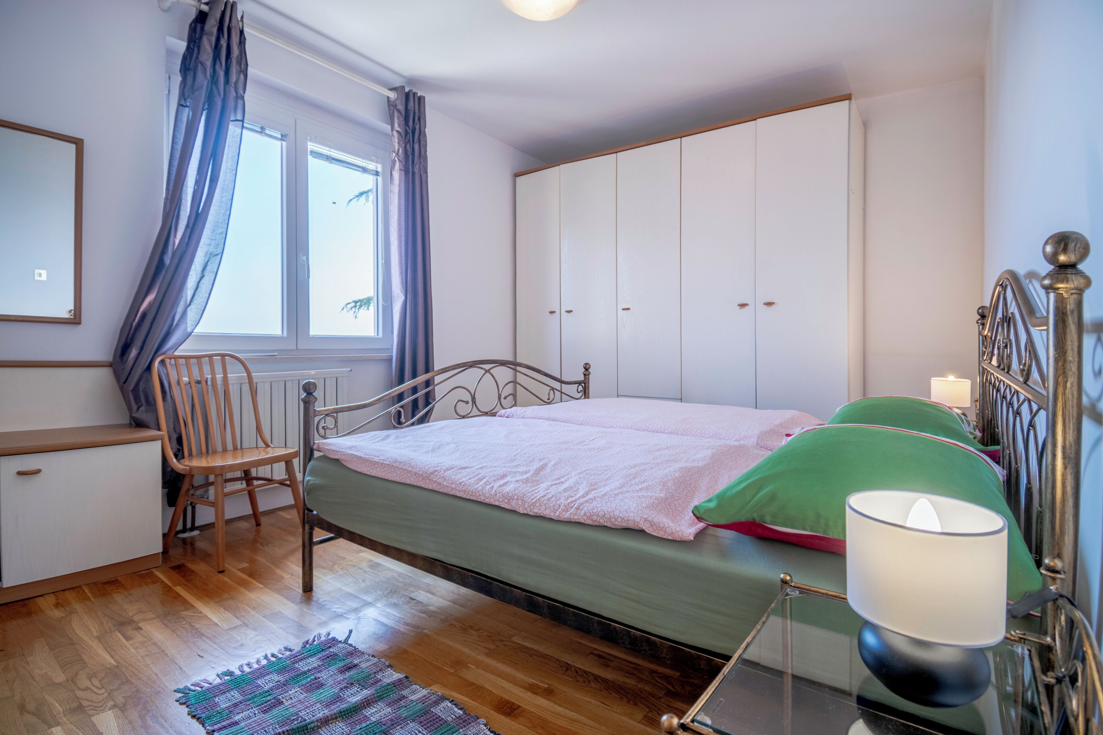 A bedroom with a bed, a chair, a wardrobe, a mirror, and a window with curtains.