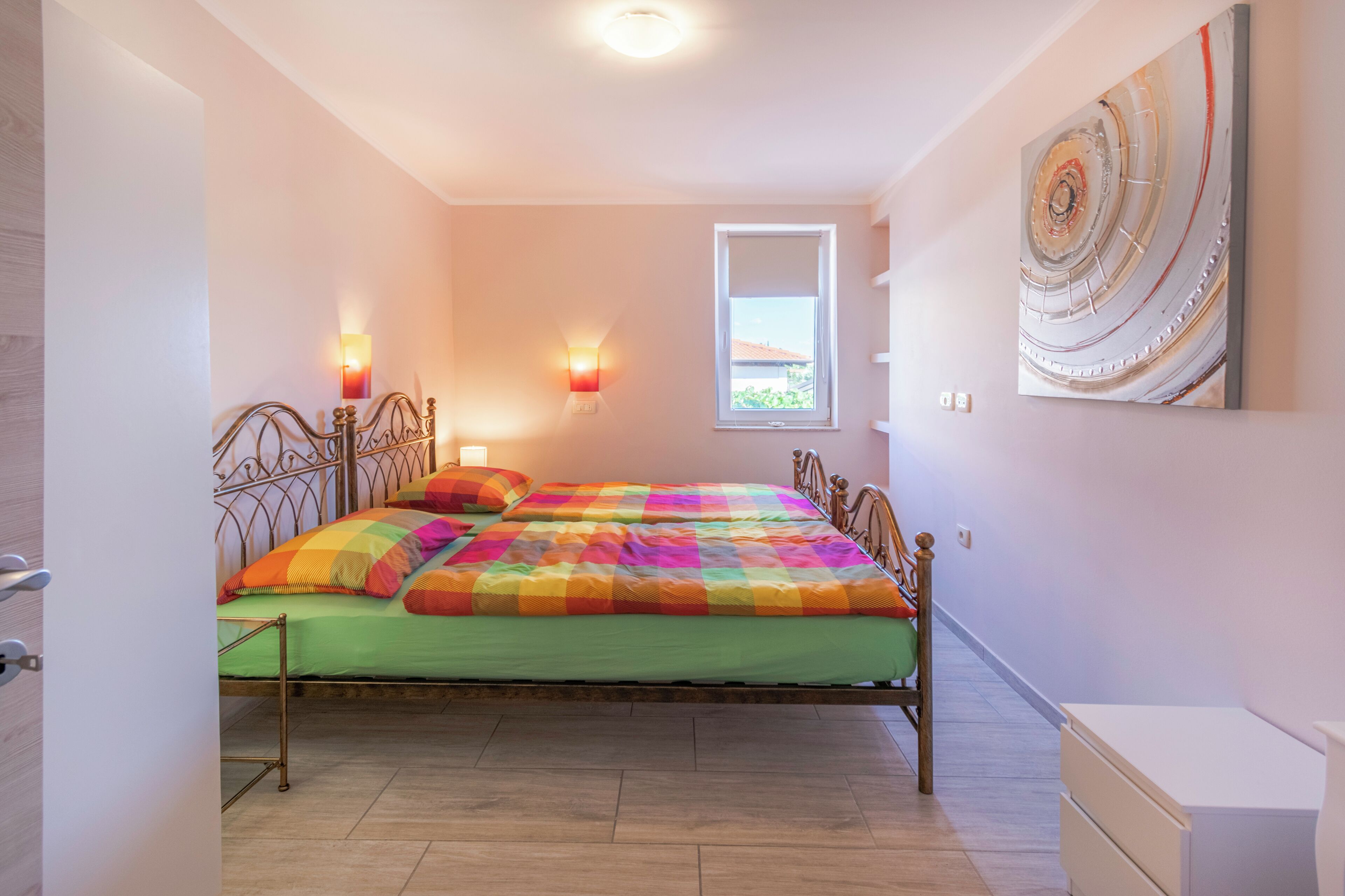 A bedroom with a double bed, two bedside lamps, a window, and a painting on the wall.