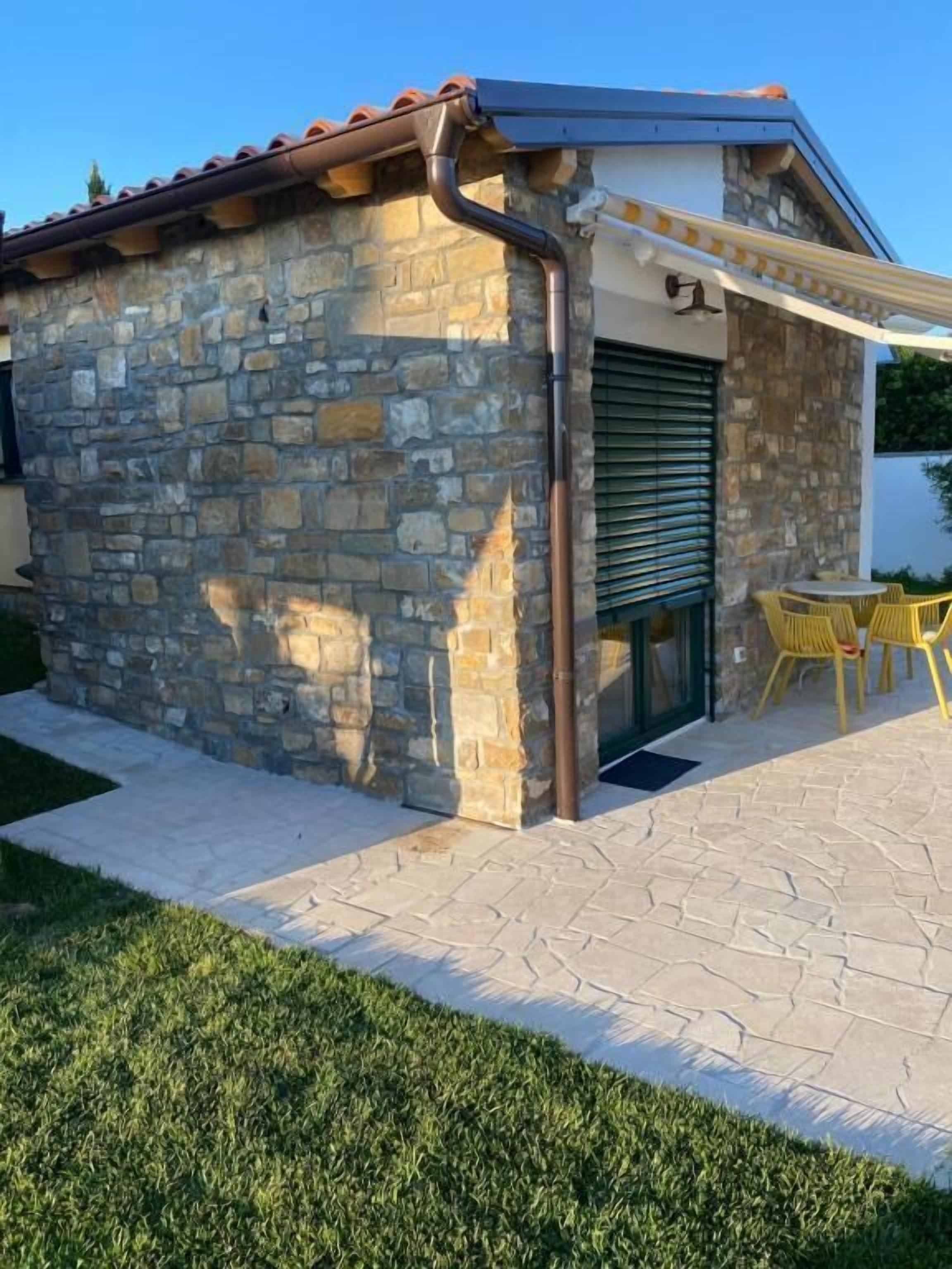 A stone building with a tiled roof, a door, and a small patio area with yellow chairs.