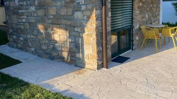 A stone building with a tiled roof, a door, and a small patio area with yellow chairs.