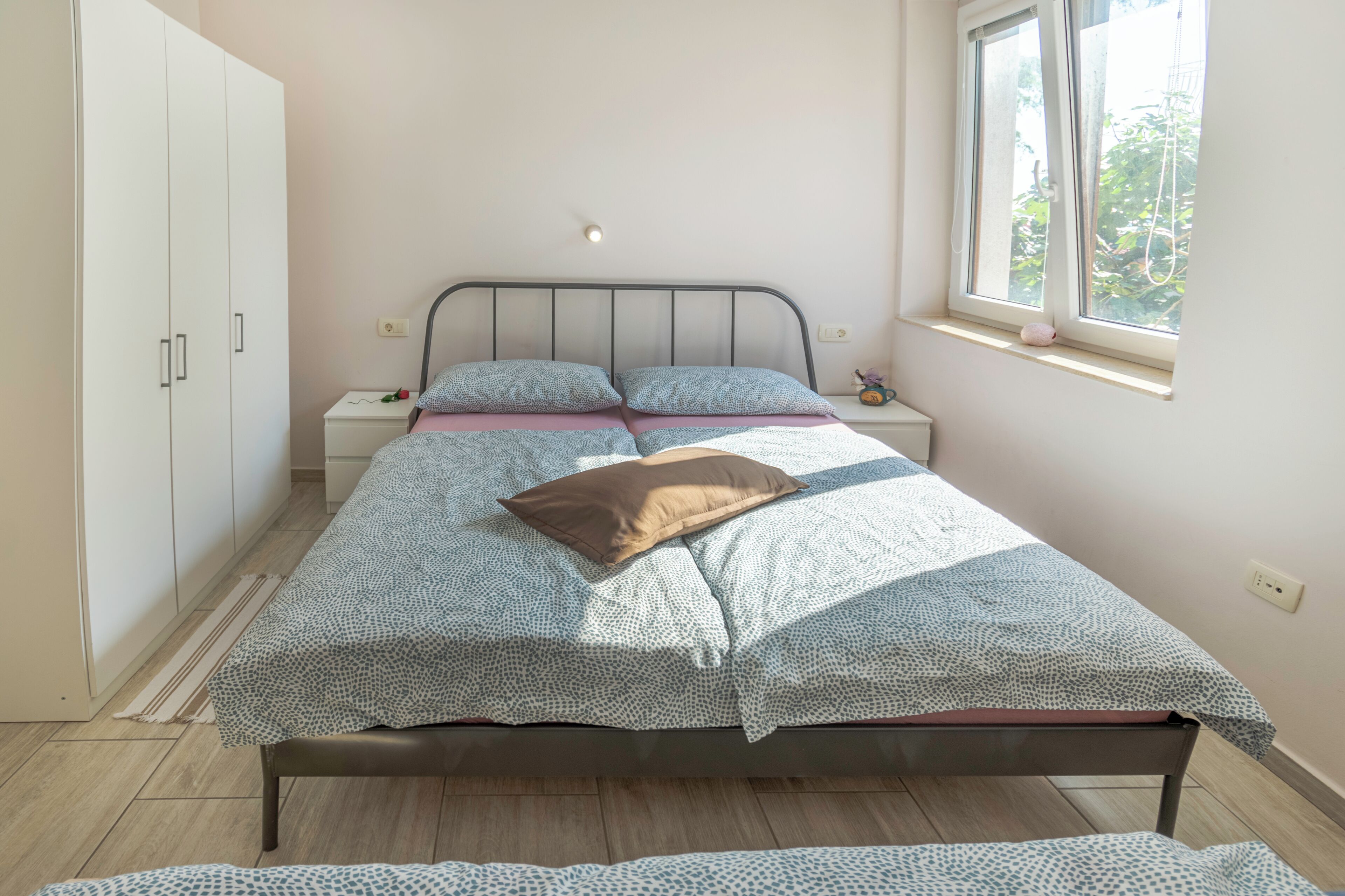 A bedroom with a bed, a nightstand, a wardrobe, and a window with a view of greenery.