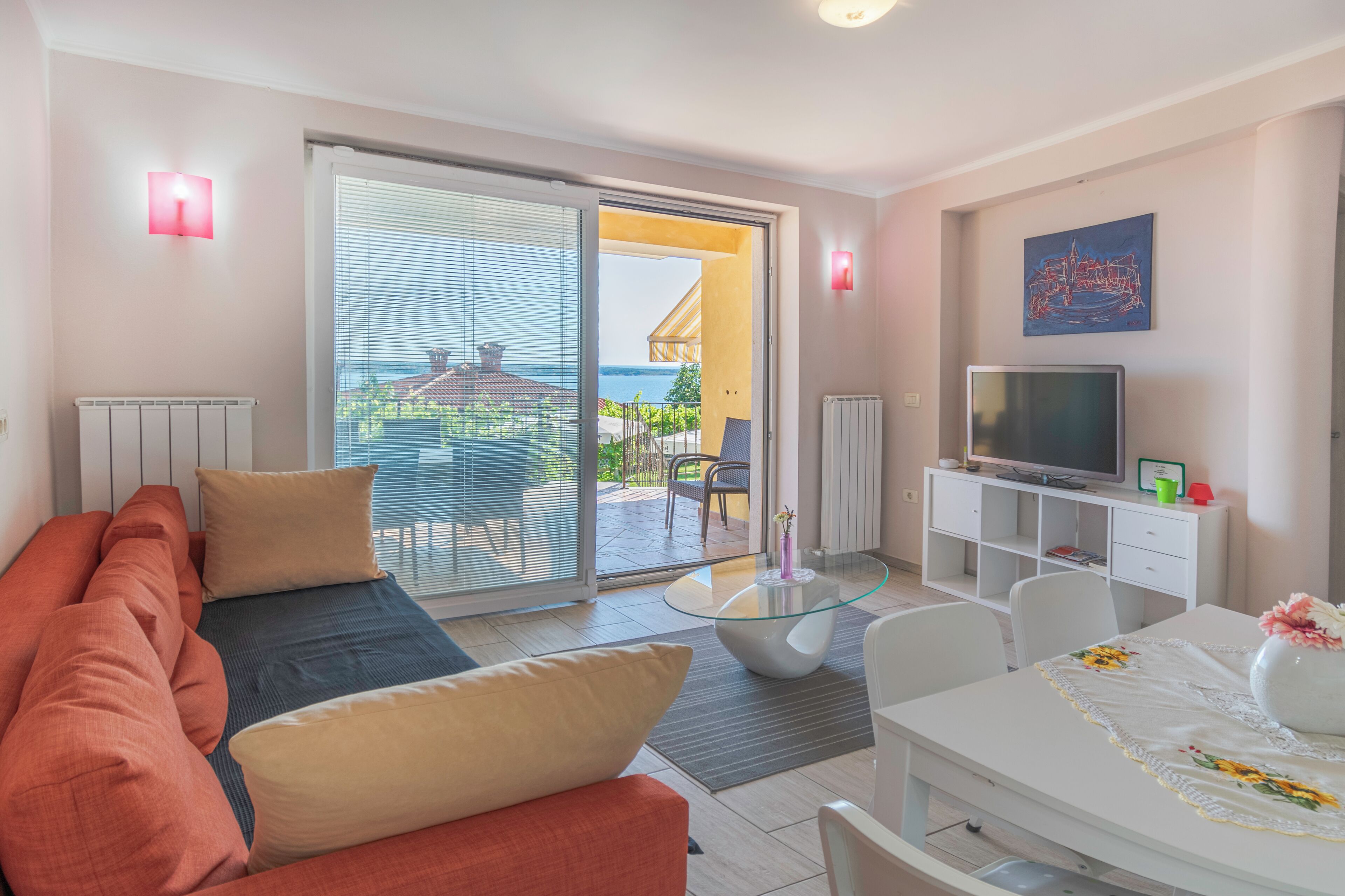 A modern living room with a sofa, a glass coffee table, a TV on a white cabinet, and a balcony with a view of the sea.
