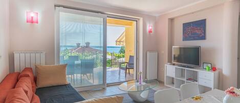 A modern living room with a sofa, a glass coffee table, a TV on a white cabinet, and a balcony with a view of the sea.