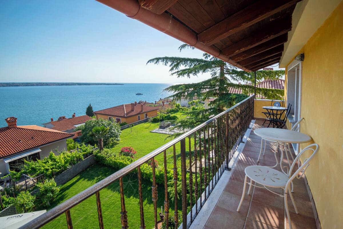 A balcony with a view of the sea, a small garden, and a table with chairs.