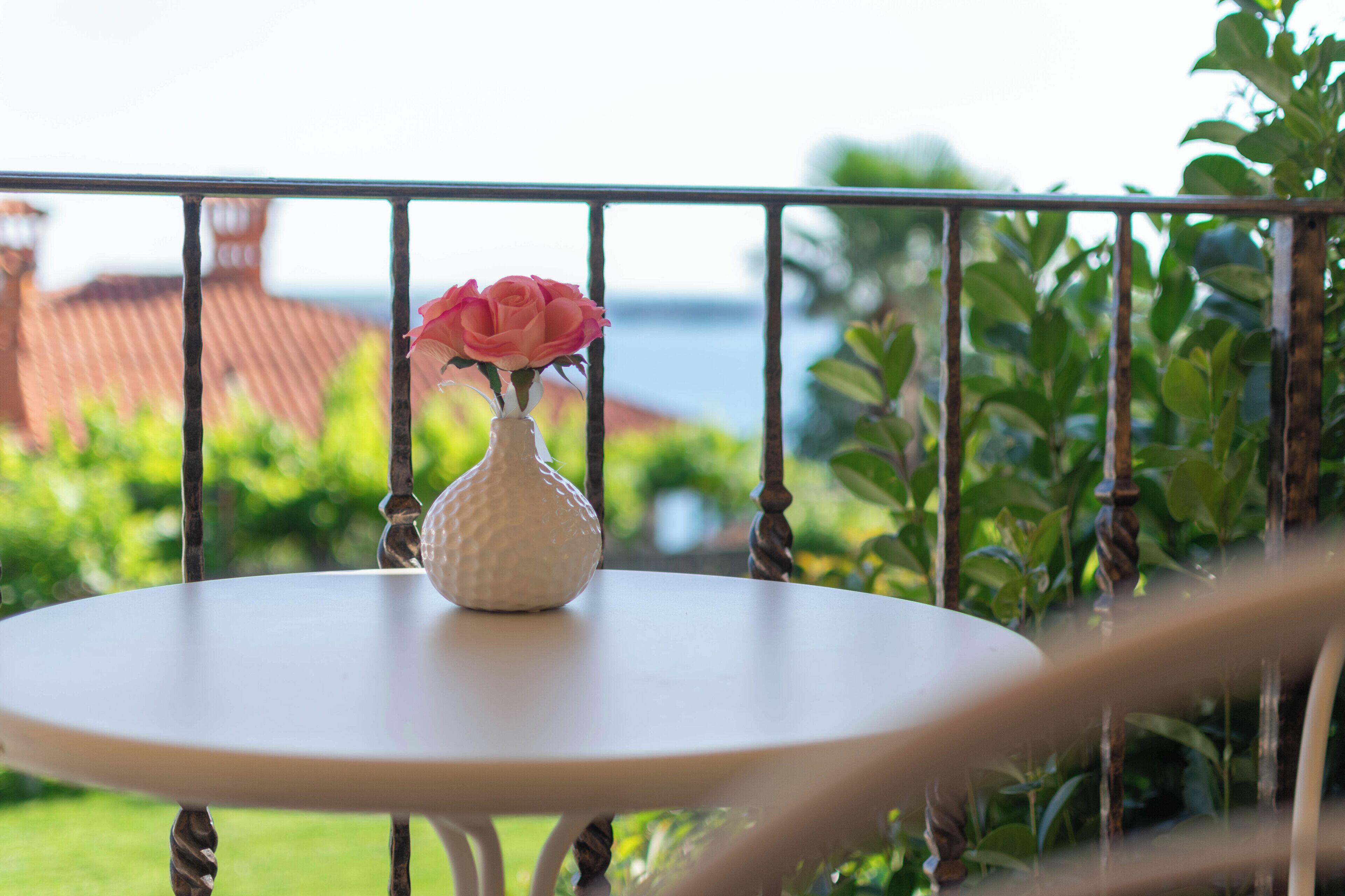 Una mesa blanca con un jarrón blanco que contiene una rosa rosa, ubicada en un balcón con vistas a la vegetación y un edificio de techo rojo.