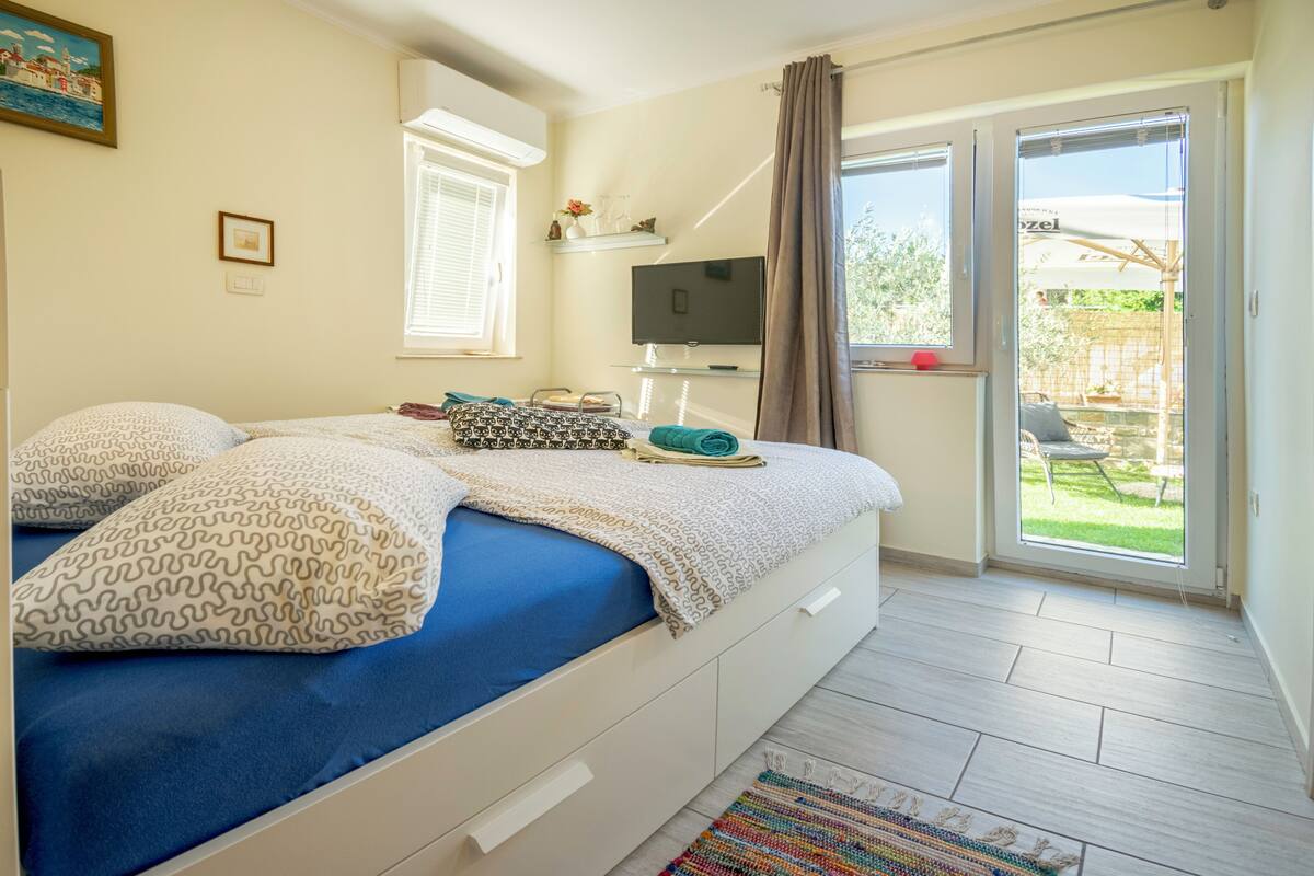 A bedroom with a bed, a TV, a window, and a view of the outdoors.