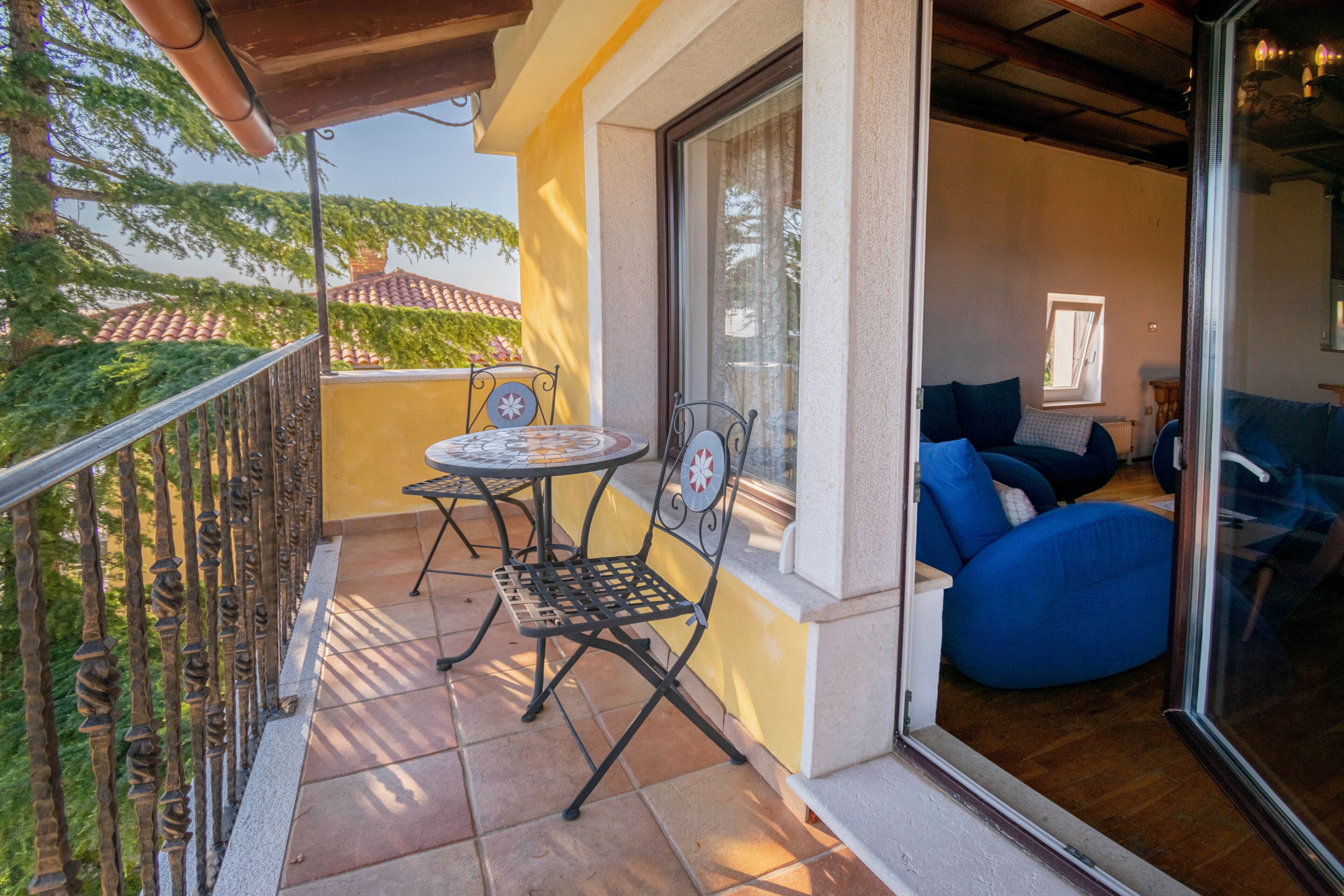 A balcony with a table and chairs, a blue sofa, and a view of a red-tiled roof and greenery.