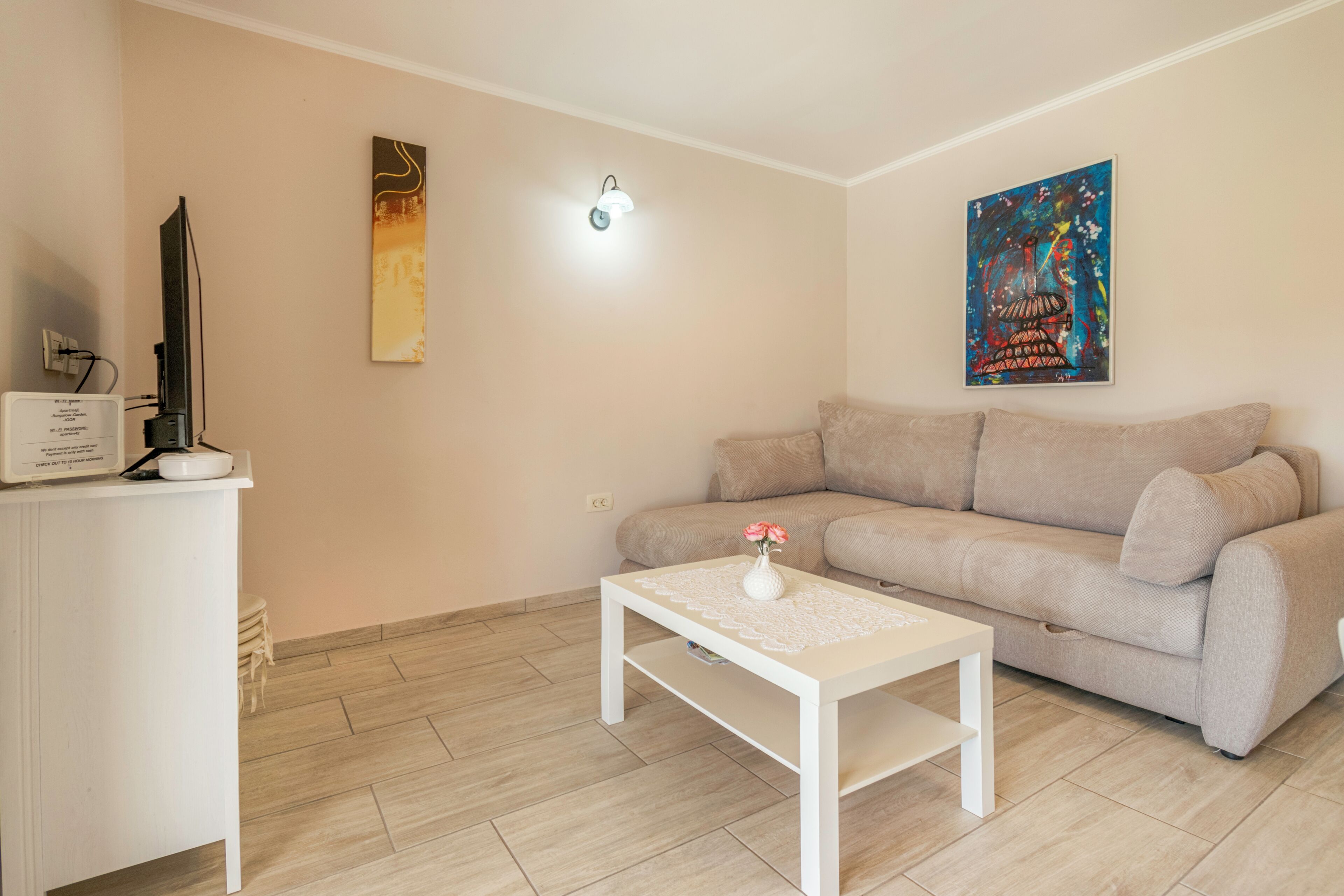 A living room with a beige sofa, a white coffee table with a vase, a flat-screen TV on a white cabinet, and two wall-mounted artworks.