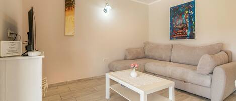 A living room with a beige sofa, a white coffee table with a vase, a flat-screen TV on a white cabinet, and two wall-mounted artworks.