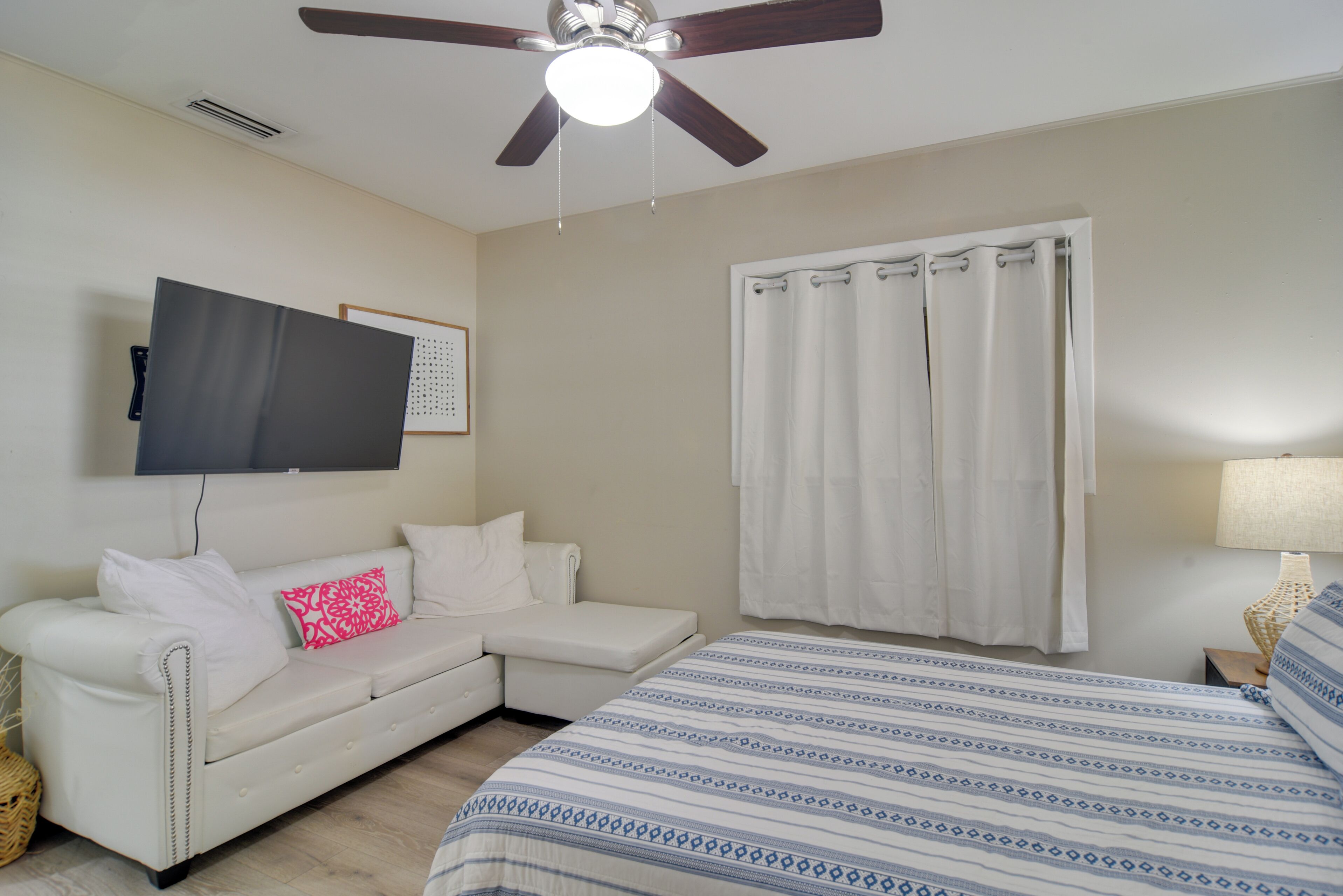 Une chambre à coucher avec un lit, un canapé blanc, un ventilateur de plafond, un téléviseur fixé au mur et une fenêtre avec des rideaux.