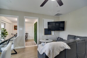 A modern living room with a grey sofa, a flat-screen TV mounted on the wall, a small round table with a plant, and a ceiling fan.