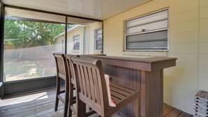 A covered outdoor seating area with wooden chairs and a bench, a wooden table, and a window with blinds. - 13 Mi to Busch Gardens: Tampa Gem w/ Grill & Yard (Tampa)