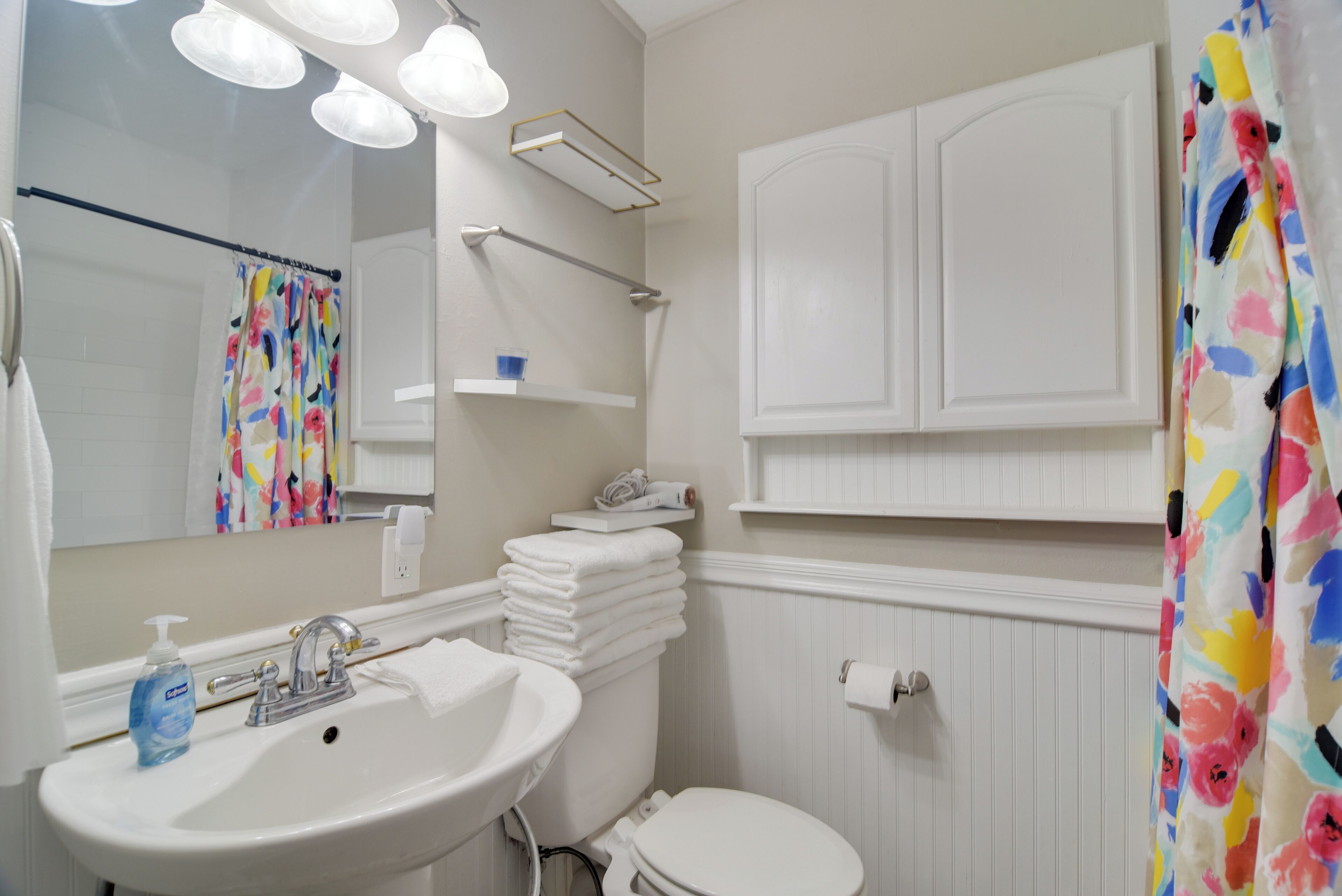 Een badkamer met een witte wastafel, spiegel en toilet. Handdoeken liggen op een plank gestapeld, en er is een douchegordijn met een bloemenmotief.