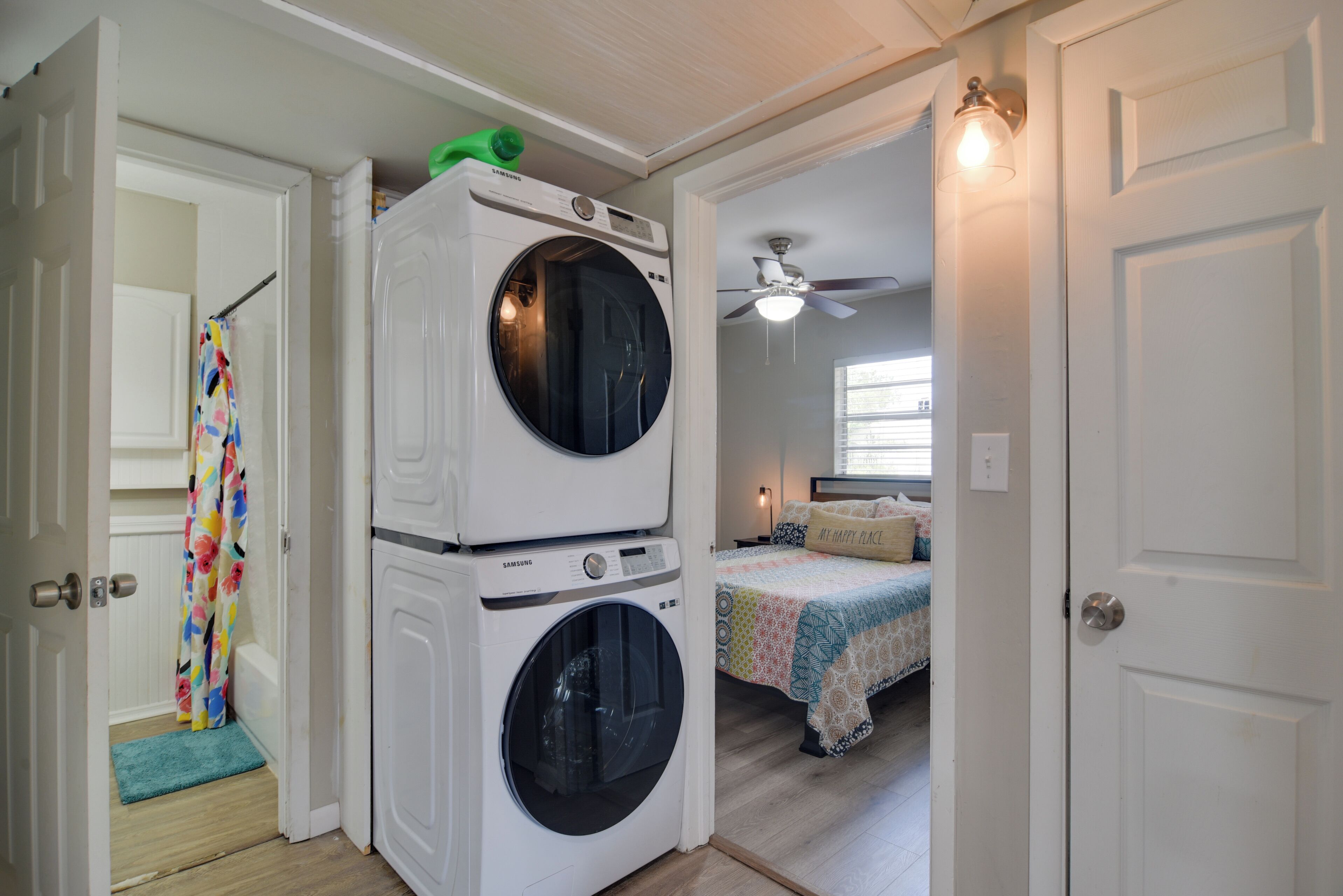 A laundry room with stacked washers and dryers, a ceiling fan, and a bedroom with a bed and window.