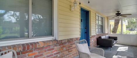 A covered porch with a brick wall, a blue door, and a seating area with wicker chairs and a round table.