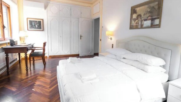 A bedroom with a large bed, a desk, and a chair. There is a wardrobe and a painting on the wall.
