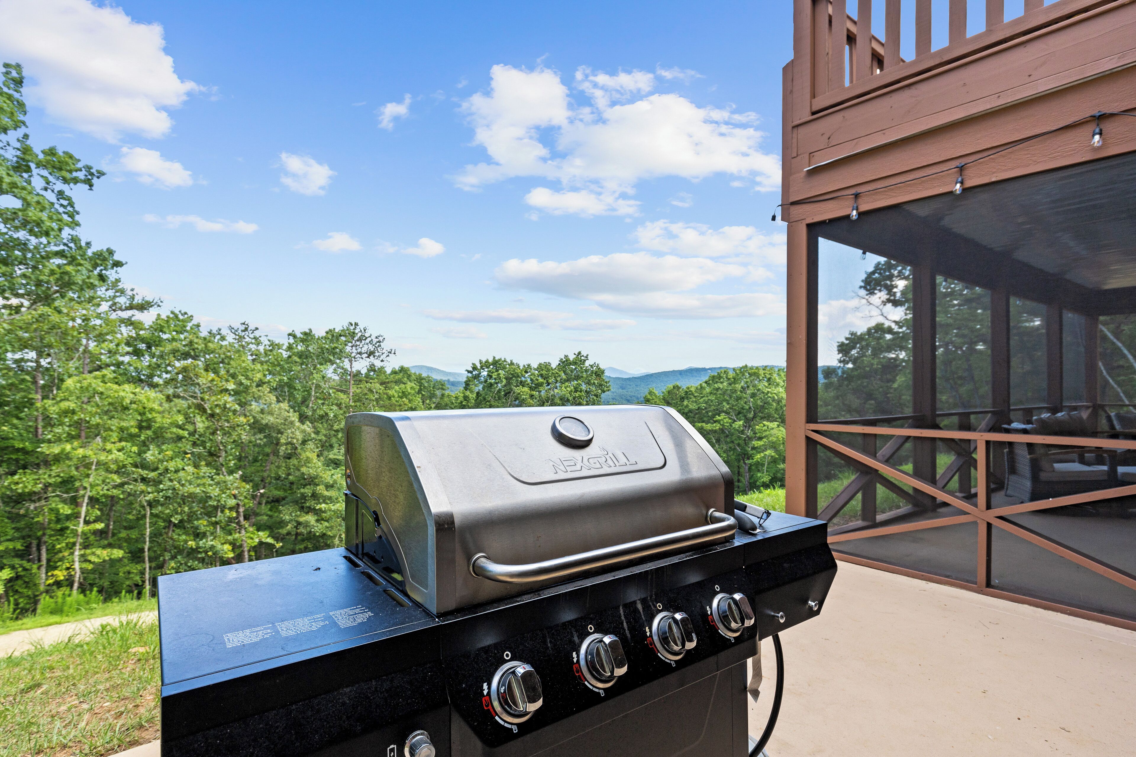 A barbecue grill with four burners, located outdoors with a view of a forest and mountains in the background.