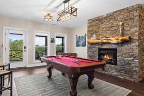 A pool table in a room with a stone fireplace, wooden flooring, and a view through a door. - Mtn View & Game Room in Morganton: Spacious Cabin! (Morganton)