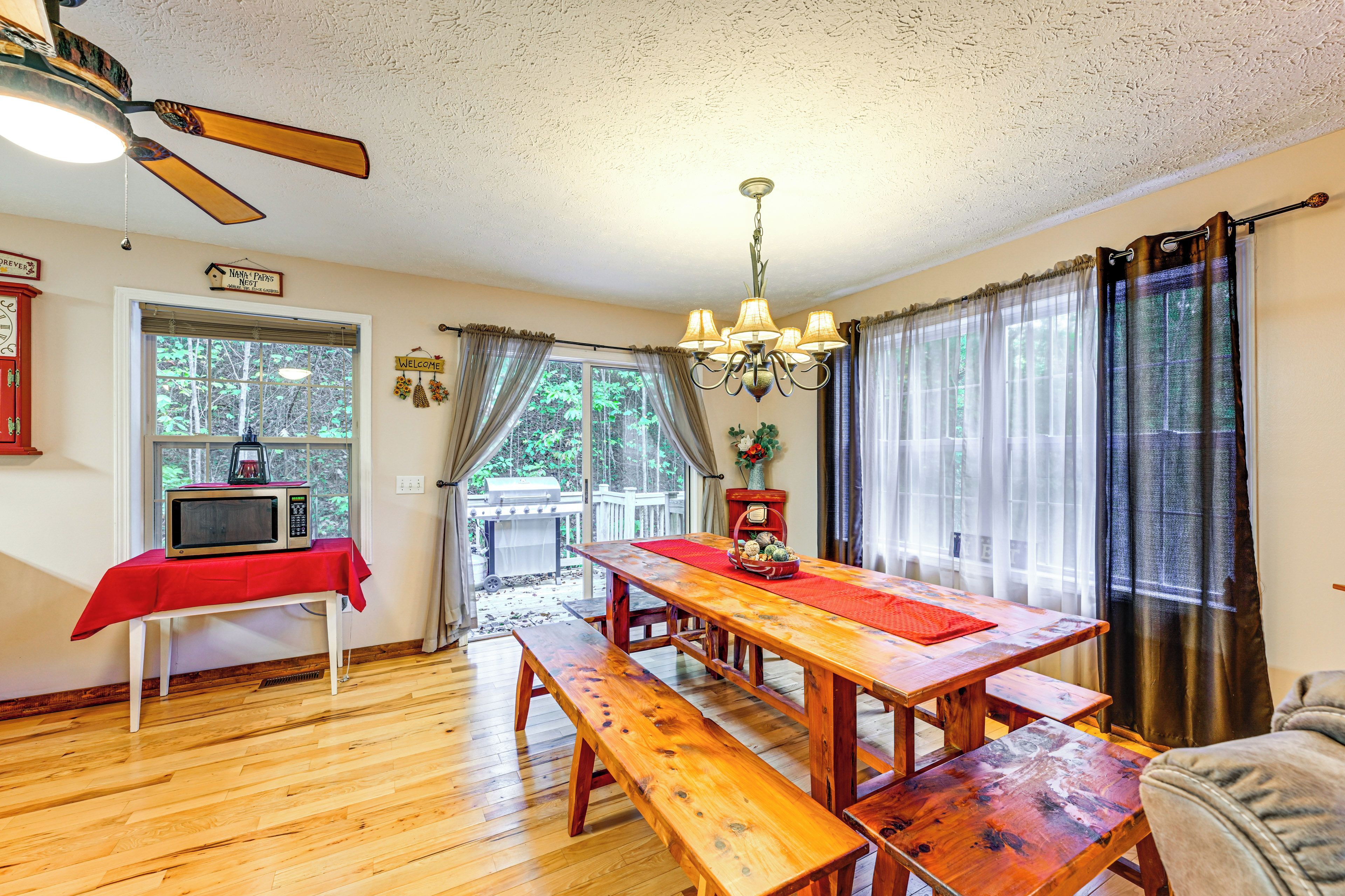 Un espace repas comprenant une table en bois, des bancs, un ventilateur de plafond et une vue sur l’extérieur grâce à de grandes fenêtres.