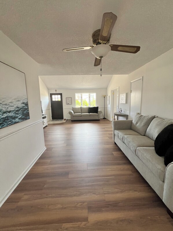 A spacious living room with hardwood flooring, a ceiling fan, a sofa, and a large window. - CHARMING NEWLY RENOVATED 4-BEDROOM HOUSE ON LAGOON IN BEACH HAVEN WEST (Stafford Township)