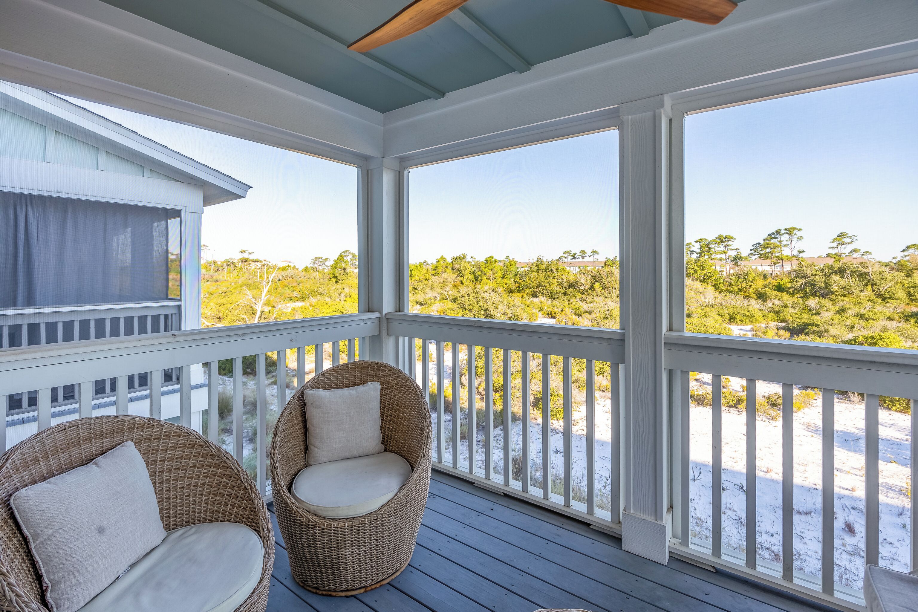 A covered porch with two wicker chairs and a view of a beach and trees.