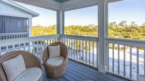 A covered porch with two wicker chairs and a view of a beach and trees.