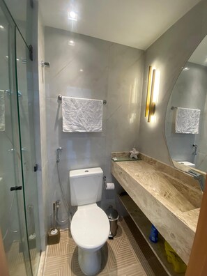 A modern bathroom with a glass shower enclosure, a toilet, a sink with a marble countertop, and a large mirror. - Villas do Porto - 301 by La Playa (Ipojuca)