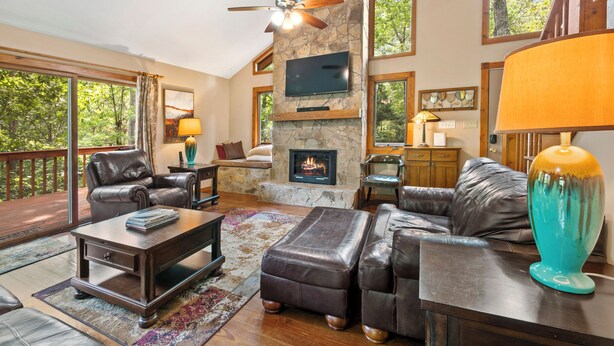 A spacious living room with a stone fireplace, leather furniture, and large windows offering a view of the outdoors. - Azure Springs- Secluded Mountain Retreat in Dahlonega. Perfect for Families! (Dahlonega)