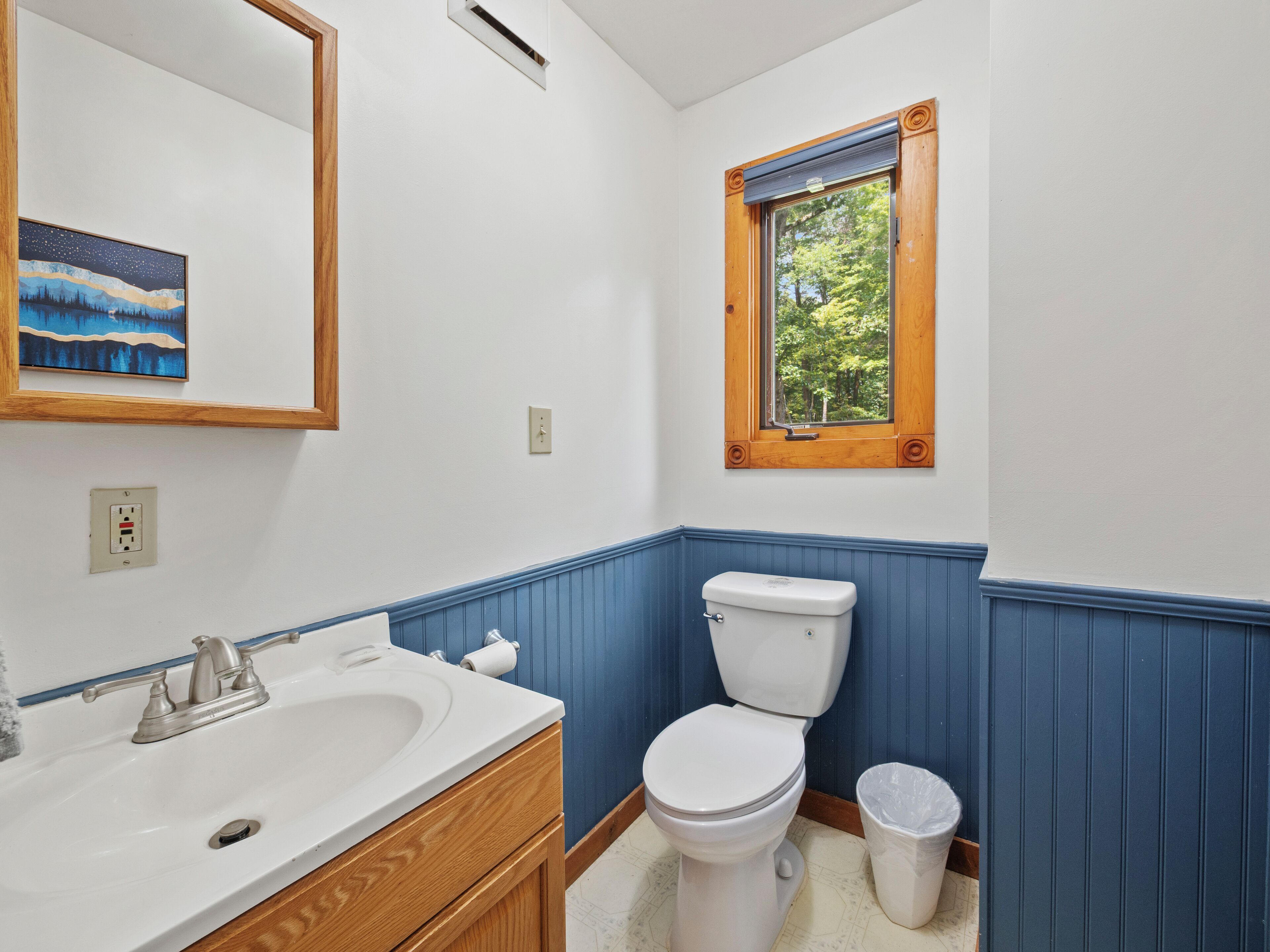 A bathroom with a white sink, wooden vanity, a white toilet, a window with a wooden frame, and a blue wainscoting.